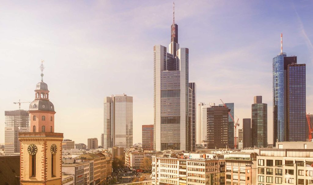 Ein Panorama von Frankfurt am Main, das die alte Uhrturm-Silhouette im Vordergrund und moderne Wolkenkratzer im Hintergrund zeigt. Die Stadtlandschaft wird von warmen, sanften Farben und einem klaren Himmel geprägt, was einen harmonischen Kontrast zwischen traditioneller Architektur und zeitgenössischem Design schafft.