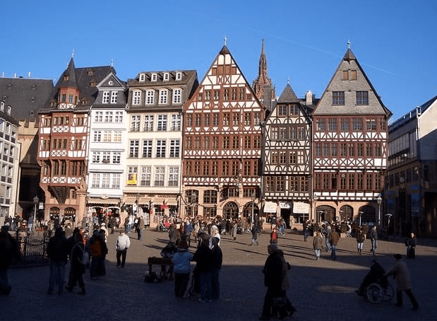 Blick auf den Römerberg in Frankfurt am Main, umgeben von historischen Fachwerkhäusern mit farbenfrohen Fassaden. Menschen schlendern über den Platz, während die Sonne hell am klaren blauen Himmel scheint. Im Hintergrund ist der markante Turm der Alten Nikolaikirche sichtbar, was die charmante Atmosphäre dieses lebhaften Stadtteils unterstreicht.