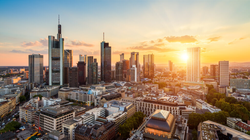 Panoramablick auf die Skyline von Frankfurt am Main bei Sonnenuntergang. Hochhäuser und Wolkenkratzer dominieren die Stadtlandschaft, während die untergehende Sonne den Himmel in warmen Orange- und Gelbtönen erleuchtet. Die Kombination aus moderner Architektur und historischen Gebäuden bildet das Stadtbild.