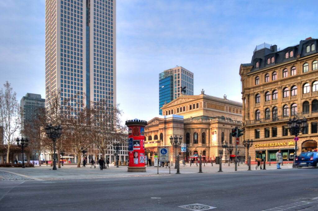 Der Opernplatz in Frankfurt am Main mit der Alten Oper im Hintergrund, umgeben von modernen Hochhäusern und historischen Gebäuden. Eine Litfaßsäule im Vordergrund ist mit Plakaten bedeckt, und Passanten überqueren den Platz. Die Szene zeigt eine Mischung aus traditioneller und moderner Architektur an einem klaren, kühlen Tag.