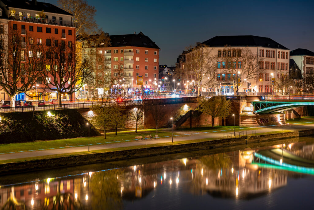 Nachtaufnahme einer Flussuferstraße in Frankfurt am Main. Die beleuchteten Gebäude und Bäume spiegeln sich im ruhigen Wasser des Flusses. Straßenlaternen und bunte Lichter beleuchten die Szene, während Autos und Fußgänger die nächtliche Stadt beleben. Eine Brücke führt über den Fluss, und die Atmosphäre ist ruhig und stimmungsvoll.