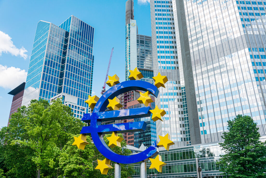 Große Euro-Skulptur mit gelben Sternen vor modernen Hochhäusern im Bankenviertel von Frankfurt am Main. Der blaue Euro-Symbol wird von glänzenden Glasfassaden und grünen Bäumen umgeben. Der Himmel ist klar und sonnig, und das Bild zeigt die Verbindung von Natur und moderner Architektur in einer der wichtigsten Finanzmetropolen Europas.