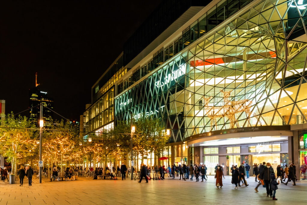 Die Einkaufsstraße Zeil in Frankfurt am Main bei Nacht, beleuchtet von Straßenlaternen und Schaufensterlichtern. Das moderne Einkaufszentrum MyZeil mit seiner markanten Glasfassade ist prominent im Bild. Menschen flanieren entlang der Straße, während die Bäume in der Fußgängerzone mit Lichterketten geschmückt sind. Im Hintergrund ist ein beleuchteter Wolkenkratzer zu sehen, der die Skyline von Frankfurt ergänzt. Die Szene zeigt eine lebendige Atmosphäre in der Stadt bei Nacht.