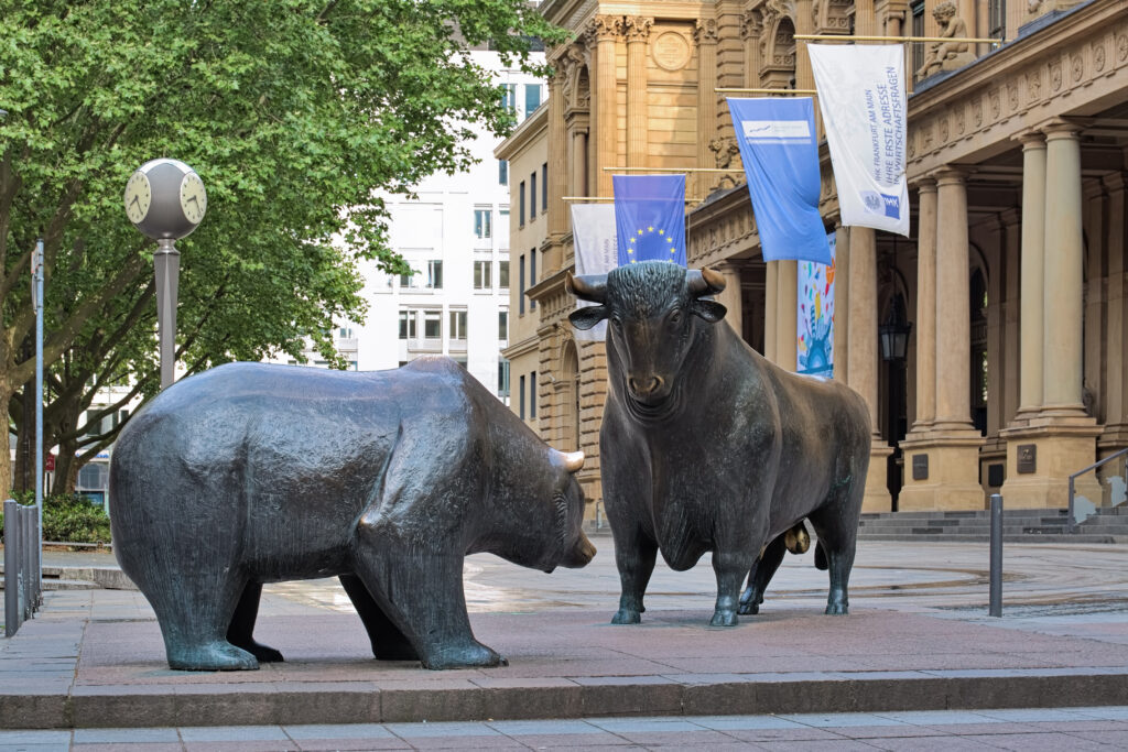 Bronzestatue von Bulle und Bär vor der Frankfurter Wertpapierbörse. Die beiden Statuen symbolisieren den Finanzmarkt und stehen sich gegenüber. Im Hintergrund sind das Gebäude der Börse sowie Bäume und europäische Flaggen zu sehen. Die Szene findet an einem sonnigen Tag statt, und die Uhr im Hintergrund zeigt die Zeit an.