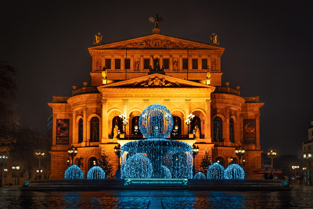 Alt-Tag: Die Alte Oper in Frankfurt am Main bei Nacht, festlich beleuchtet in warmem Orange. Vor dem Gebäude steht ein Brunnen, der mit blauen Lichterketten dekoriert ist, die weihnachtliche Stimmung verbreiten. Weihnachtsbäume und Straßenlaternen ergänzen die festliche Szene, während die Architektur der Alten Oper im Hintergrund eindrucksvoll hervorsticht. Ein Schild vor dem Brunnen trägt den Schriftzug „Season's greetings and best wishes for the new year.“