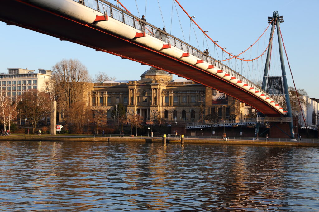 Blick auf das Städel Museum in Frankfurt am Main, das hinter der Holbeinsteg-Fußgängerbrücke am Ufer des Mains liegt. Die Brücke spannt sich im Vordergrund über den Fluss, und das ruhige Wasser reflektiert die goldene Abendsonne. Menschen spazieren entlang des Ufers, und die Szenerie vermittelt eine ruhige, spätnachmittägliche Atmosphäre.