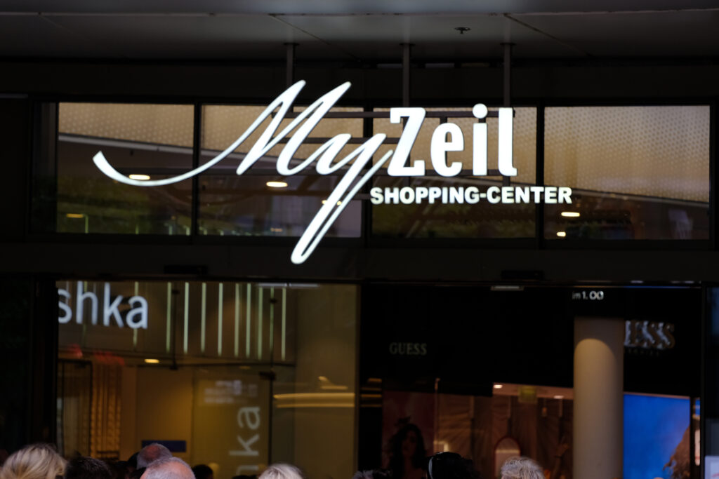 Leuchtendes Logo des MyZeil Shopping-Centers in Frankfurt am Main, aufgenommen am Eingang des Einkaufszentrums. Der Schriftzug „MyZeil“ in geschwungener Schrift und „Shopping-Center“ in Blockbuchstaben sind prominent zu sehen. Im Hintergrund sind Schaufenster und Menschen zu erkennen, die sich im Eingangsbereich versammeln. Die Szene zeigt das geschäftige Treiben im Herzen der Einkaufsmeile Zeil.